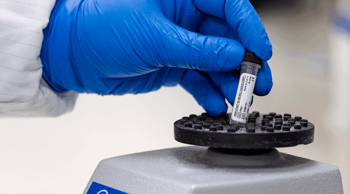 A lab technician wearing blue gloves holds a vial of Streck MDx-Chex for BCID2, a molecular quality control for sepsis testing, over a lab vortex mixer. This image illustrates the use of the product in a clinical or research laboratory setting.
